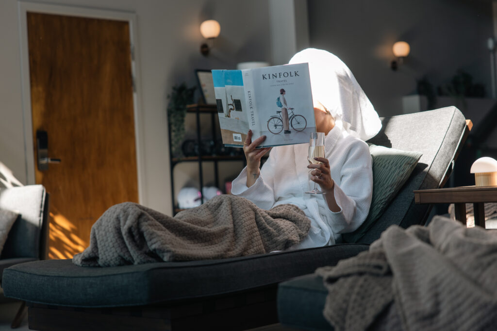 Gäst i badrock läser tidning med glas champagne i modern hotellsvit med naturgröna textilier