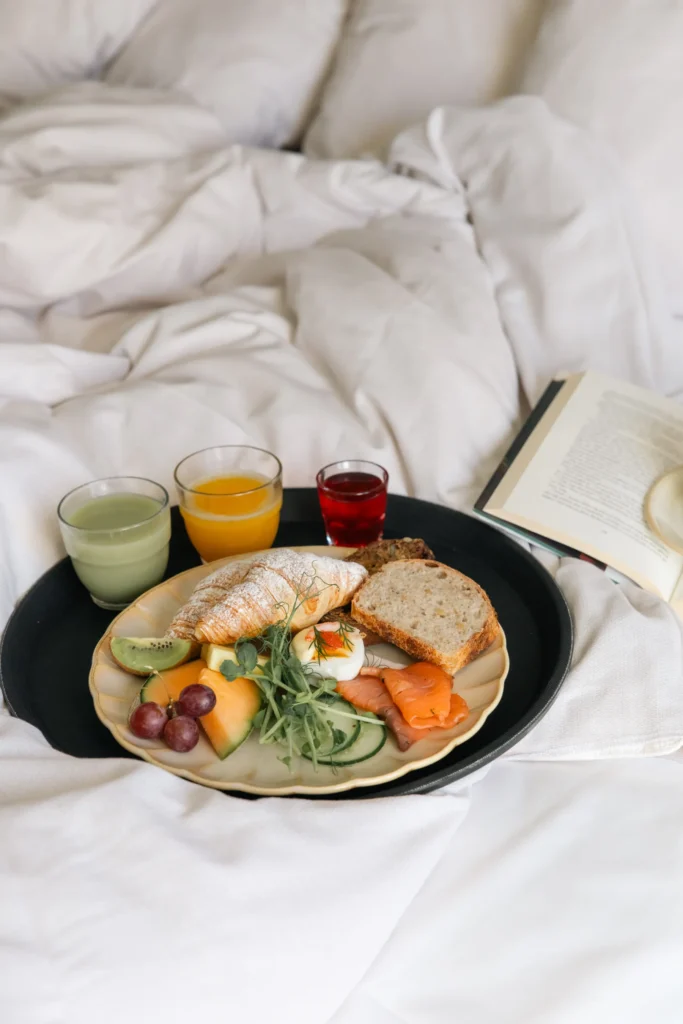 Frühstückstablett mit Croissant, Eiern, Lachs, Obst und Saft, das auf dem Bett serviert wird und neben dem ein aufgeschlagenes Buch liegt
