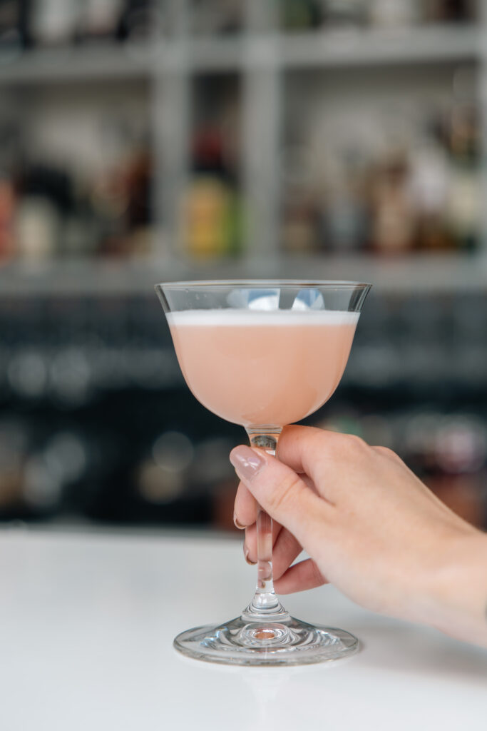 Hand som håller rosa cocktail i elegant glas framför hotellbar med flaskor i bakgrunden