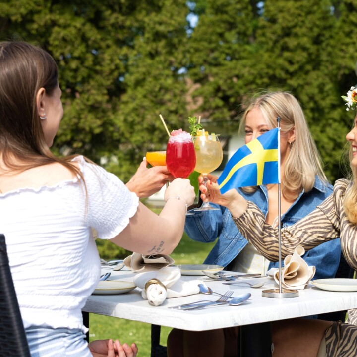 Gäster skålar med färgglada cocktails vid utomhusbord med svensk flagga i bakgrunden på solig dag