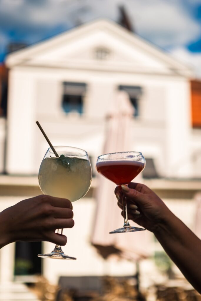 Två händer skålar med cocktails framför vit villa vid Vättern - perfekt för möhippa med avkoppling och glädje