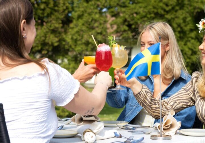 Gäster skålar med färgglada cocktails vid utomhusbord med svensk flagga i bakgrunden på solig dag